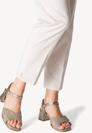 Persona vestida con pantalones cortos blancos y sandalias de tacón cuadrado metálico plateado con correas en el tobillo, de pie sobre una superficie blanca.