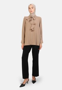 Beige blouse met ruches aan de voorkant, lange mouwen, zwarte wijde broek en glanzende zwarte pumps met spitse neuzen. Model draagt ook een bijpassende hoofddoek.