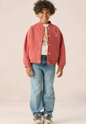 Rode katoenen jas met drukknoopjes en zijzakken, gedragen over een graphic t-shirt, gecombineerd met lichtblauwe jeans en meerkleurige sneakers.