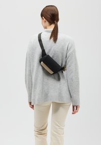 Czarna torba crossbody o gładkiej powierzchni, z złotymi akcentami, regulowanym paskiem noszona na jasnoszarym swetrze i kremowych spodniach.