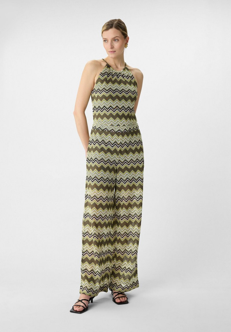 Green and patterned jumpsuit with a halter neck design, featuring zigzag stripes in black, white, and brown tones. Wide-leg silhouette.