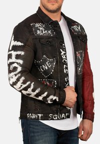 Veste en denim noir avec des graphismes peints, des clous et des patches. Accents rouges sur les manches et plusieurs designs blancs sur les bras et le corps. Fermeture à boutons.