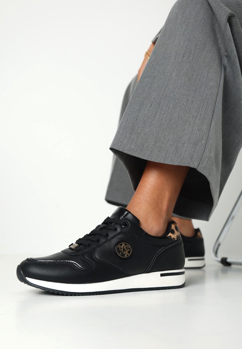 Baskets en cuir noir avec une semelle blanche, arborant un logo sur le côté et un accent en imprimé léopard au niveau du talon. Portées avec un pantalon gris.