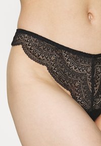 String noir en dentelle mettant en avant des détails de motif élaborés, un bord festonné et une texture douce. La moitié d'une taille et d'une hanche est visible.
