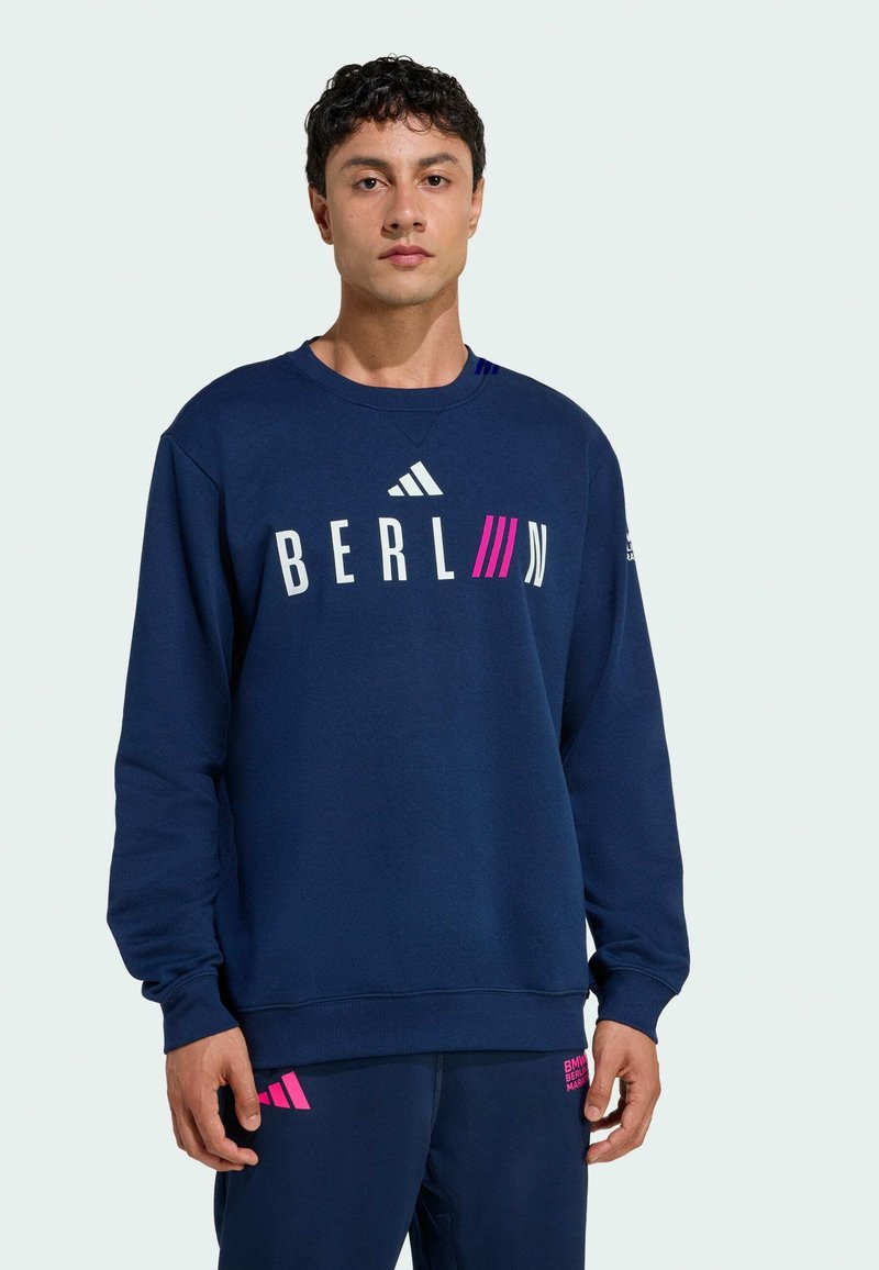 Navy blue sweatshirt with "BERLIN" printed in white and pink, featuring crew neck, long sleeves, and logo accents on shoulders.