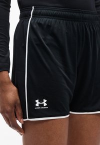 Μαύρα αθλητικά σορτς με λευκή επένδυση και λογότυπο Under Armour, φορεμένα από άτομο με σκούρο δέρμα, με το ένα χέρι να ακουμπά κοντά στο τελείωμα.