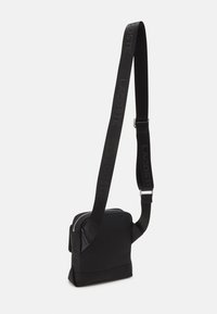 Borsa a tracolla nera realizzata in materiale liscio, con tracolla staccabile con il marchio "LACOSTE", chiusura con zip e design angolare.