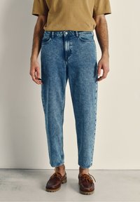 Modri denim jeans z ohlapnim krojem, izblitem pranjem, petimi žepi in gumbom za zapiranje, v paru s rjavimi vezalkami.