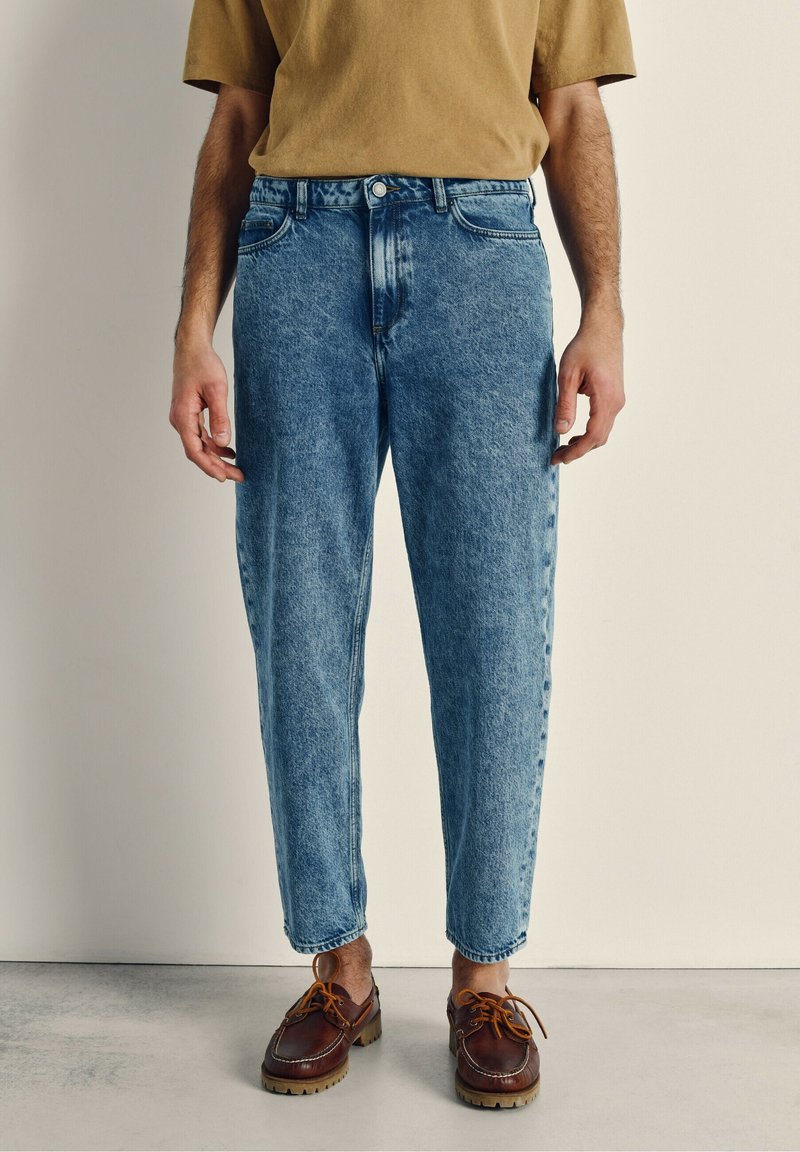 Modri denim jeans z ohlapnim krojem, izblitem pranjem, petimi žepi in gumbom za zapiranje, v paru s rjavimi vezalkami.