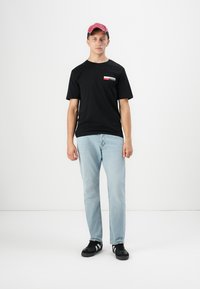 Jonge man die staat en een rode pet, een zwart Jack & Jones t-shirt, lichtblauwe spijkerbroek en zwarte sneakers draagt tegen een witte achtergrond.