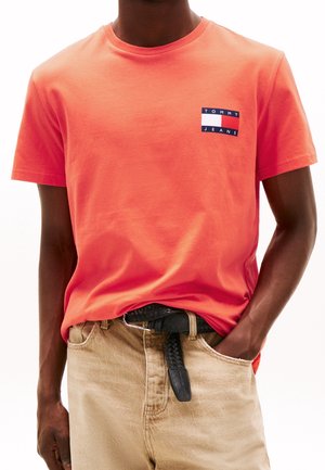 Adulto con indosso una t-shirt Tommy Jeans rosso corallo infilata in pantaloni beige con cintura nera intrecciata, mano in tasca.