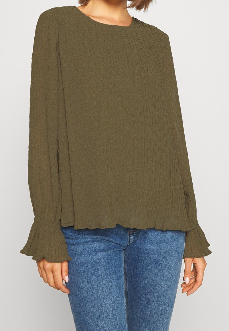 Femme portant un chemisier texturé vert olive à manches longues avec poignets évasés et jean bleu, visible du cou jusqu'aux hanches.