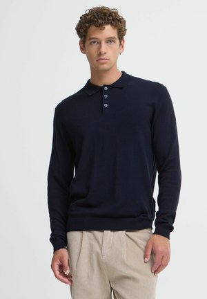 Mann mit lockigem Haar trägt ein dunkelblaues langärmeliges Poloshirt und beige Hosen und steht vor einem schlichten hellen Hintergrund.