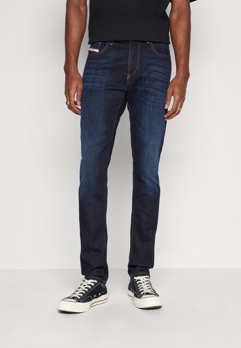 Jean en denim bleu foncé avec une coupe droite, couture contrastante, conception à cinq poches, porté avec des baskets noires et une chemise noire.