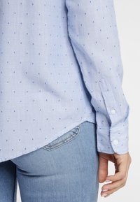 Helle blaue Langarmbluse mit kleinen dunkelblauen Punkten, ausgestattet mit einem knöpfbaren Bündchen und einem abgerundeten Saum, kombiniert mit hellblauen Jeans.