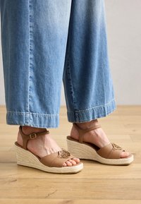 Sandales à talons compensés en cuir tan avec une semelle en jute tressé, un bout ouvert et une bride de cheville réglable. Présente un accent métallique décoratif à l'avant.