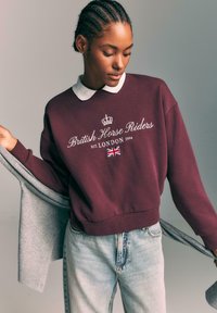 Bordo pulover s kapuco z belim napisom "Britanski konjeniški vozniki, ustanovljeni LONDONA 1994" in grafiko krone; povezan s svetlo modrimi jeans hlačami in sivim kardiganom.