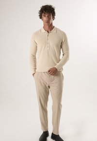 Beżowy sweter z dzianiny z kołnierzykiem i plisą na trzy guziki, zestawiony z jasnobeżowymi spodniami i czarnymi botkami za kostkę.