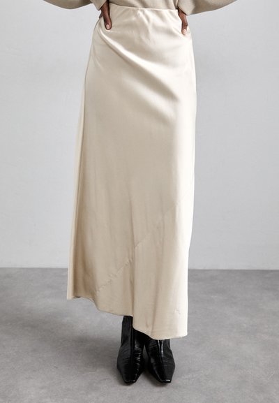 Jupe maxi en satin beige avec une coupe biaisée, affichant une texture lisse et un éclat subtil, associée à des bottes à talons noires brillantes.