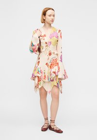 Φλοράλ φόρεμα-μανδύας σε ανοιχτό μπεζ χρώμα με μακριά μανίκια τύπου καμπάνα, βολάν στο τελείωμα και δεσίματα στη μέση. Συνδυασμένο με καφέ δερμάτινα παπούτσια με κορδόνια.