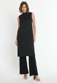 Schwarzes ärmelloses Kleid mit strukturiertem Rautendesign, hohem Kragen, knielang, getragen über schwarzen weit geschnittenen Hosen; goldene Plateau-Heels.