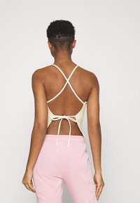 Cream tank top featuring crossed straps and a tied back, paired with pink sweatpants. The fabric appears soft with a fitted design.