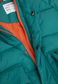 Tyrkysová péřová bunda s oranžovým podšívkou, vybavená celým zipem, patentními knoflíky a bočními kapsami. Celá bunda má prošívanou strukturu.