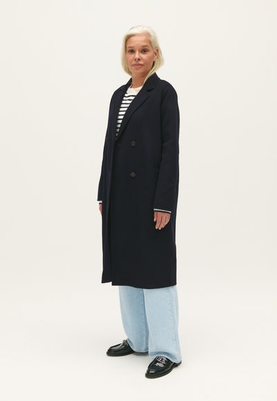Cappotto doppiopetto blu navy con tasche laterali, indossato sopra una camicia a righe bianca e nera, abbinato a jeans a gamba larga azzurri e scarpe nere.