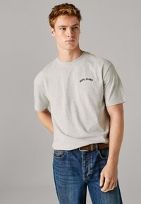 Ανοιχτό γκρι βαμβακερό t-shirt με κοντά μανίκια, διακοσμημένο με κεντητό λογότυπο "Pepe Jeans" σε ναυτικό μπλε στο στήθος, συνδυασμένο με σκούρο μπλε τζιν παντελόνι.