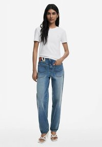 Weißes Kurzarm-T-Shirt und blaue, hoch taillierte Jeans mit entspanntem Schnitt. Weiche Textur; die Jeans haben Gesäßtaschen und farbenfrohe Akzente am Bund.