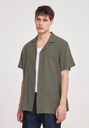 Camicia corta verde oliva in tessuto testurizzato, con bottoni al davanti e colletto a punta, indossata sopra una canotta bianca.