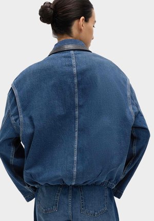 Donna con capelli scuri raccolti in uno chignon indossa una giacca di jeans oversize blu e jeans, vista da dietro su uno sfondo chiaro e semplice.