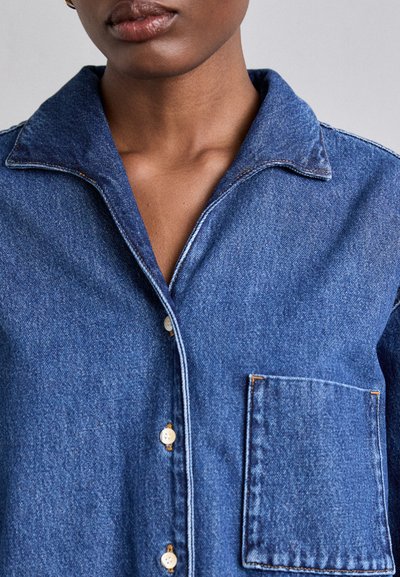 Blue denim shirt with a pointed collar, front buttons, and a patch pocket. Features contrasting stitching and textured fabric.
