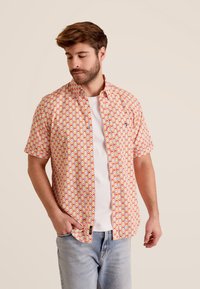 Korte mouwen shirt in oranje, met een geometrisch patroon in blauw en rood. Knop-down kraag, ontspannen pasvorm, gedragen over een wit T-shirt.