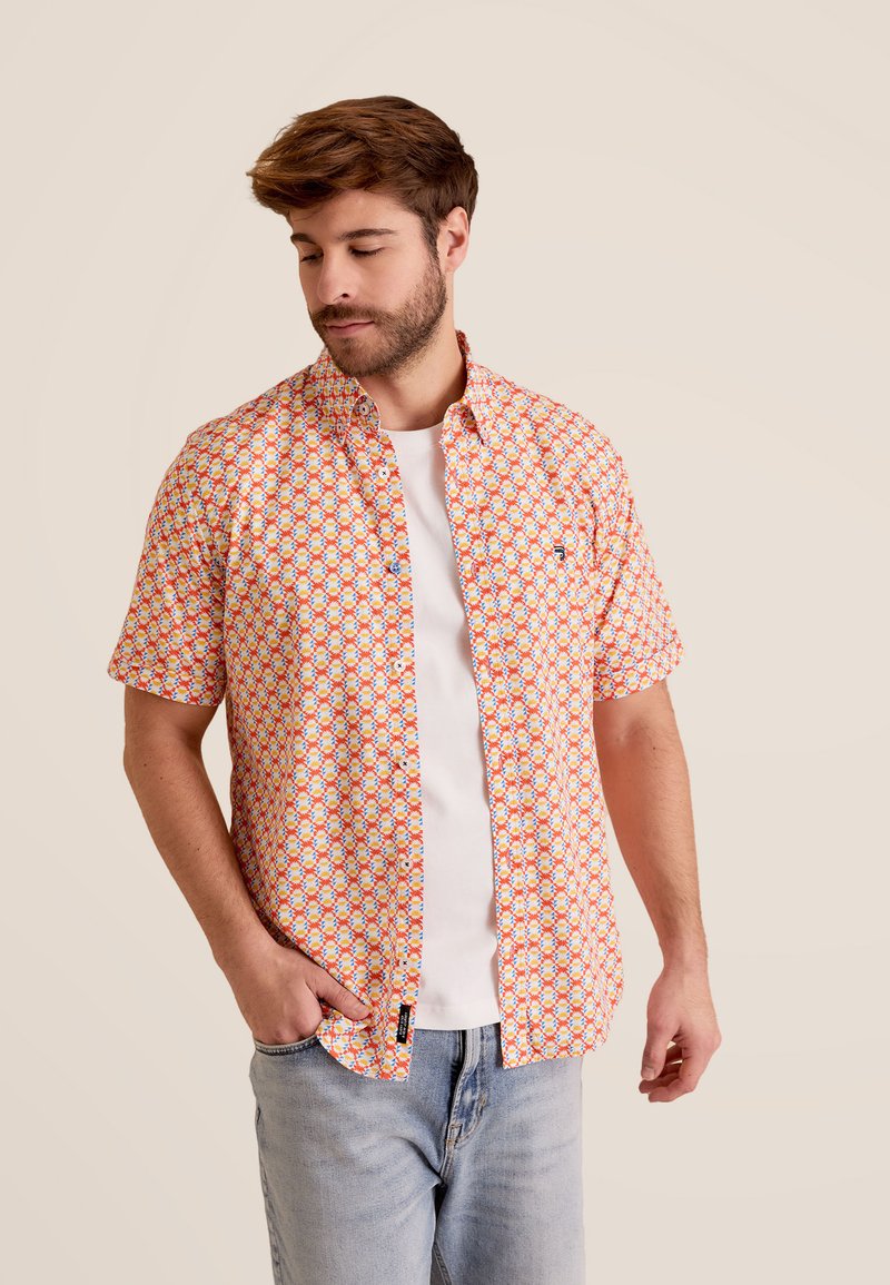 Korte mouwen shirt in oranje, met een geometrisch patroon in blauw en rood. Knop-down kraag, ontspannen pasvorm, gedragen over een wit T-shirt.