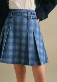 Džínová sukně s modrým károvaným vzorem, sklady a dvojitým přezkovým páskem. Materiál má jemný, texturovaný povrch.