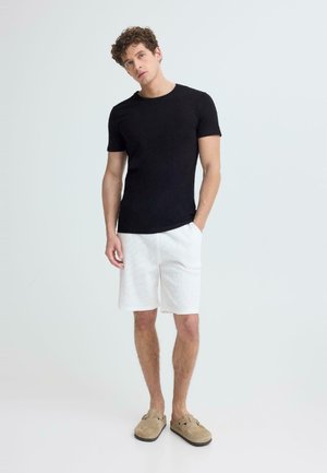 Mlad moški stoji z eno roko v žepu, oblečen v črno majico, bele kratke hlače in bež natikače, na enostavni svetli podlagi.