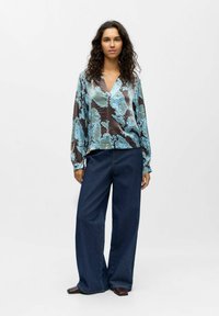 Silky, patterned blouse in shades of blue and brown, paired with wide-legged dark denim jeans. The blouse has a V-neck and long sleeves.