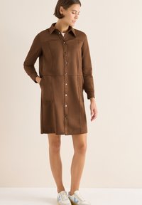 Abito marrone con bottoni, maniche lunghe, due tasche frontali e colletto, realizzato in tessuto morbido con dettagli di cucitura visibili. Indossato con sneakers bianche.