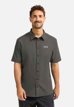 Uomo sorridente che indossa una camicia a maniche corte grigio scuro con taschino sul petto e logo Jack Wolfskin sul lato sinistro.