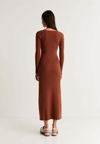 Das gerippte, langärmelige, braune Kleid hat eine taillierte Silhouette und einen runden Ausschnitt. Es wird mit klobigen, offenen Plateauabsätzen getragen.