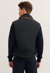 Dunkelgraue Weste für Herren mit hohem Kragen, kontrastierendem braunen Akzent am Hals und navy-blauen Rippärmeln. Glatte Textur, taillierte Passform.