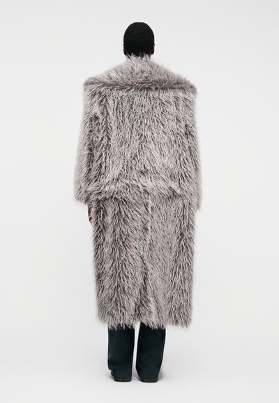 Personne portant un long manteau en fourrure grise ébouriffée, debout de dos sur un fond blanc uni.