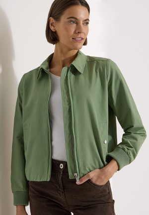 Femme portant une veste zippée vert clair avec des poignets côtelés, un haut blanc et un pantalon foncé, regardant sur le côté avec une main dans la poche.