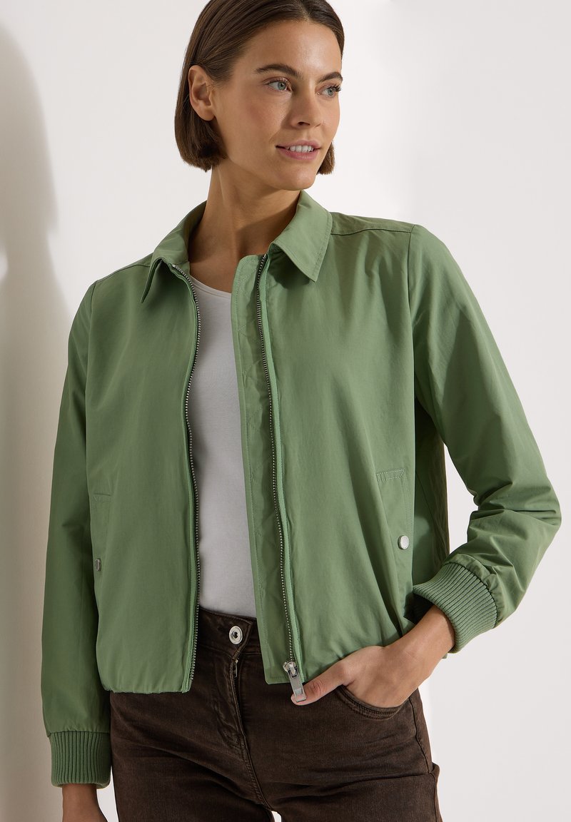 Femme portant une veste zippée vert clair avec des poignets côtelés, un haut blanc et un pantalon foncé, regardant sur le côté avec une main dans la poche.