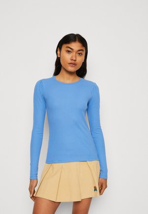 Maglia a maniche lunghe a costine di colore azzurro chiaro, con scollo a giro e maniche leggermente puffate, abbinata a una gonna beige plissè.