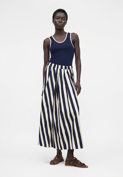 Γυναίκα όρθια φοράει μπλε σκούρο αμάνικο τοπ, ψηλόμεσο πλατύ παντελόνι με κάθετες ρίγες σε μπλε σκούρο και λευκό και καφέ σανδάλια.