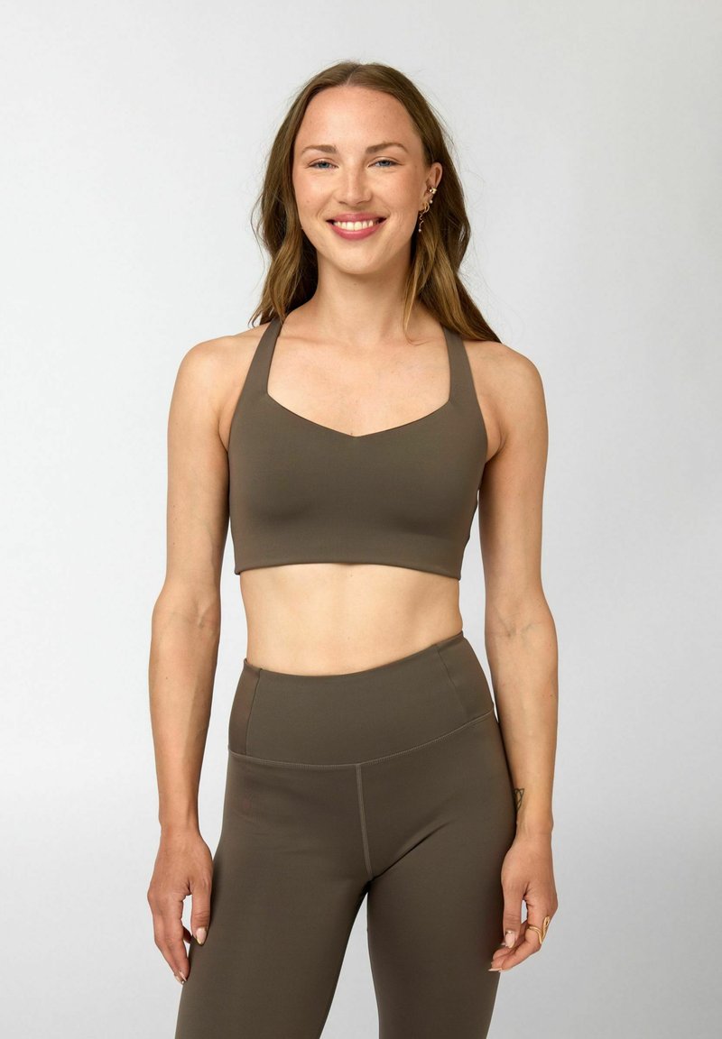 Jonge vrouw die glimlacht, gekleed in een bijpassende olijfgroene sportbeha en eenlegging met hoge taille, staand tegen een lichtgrijze achtergrond.