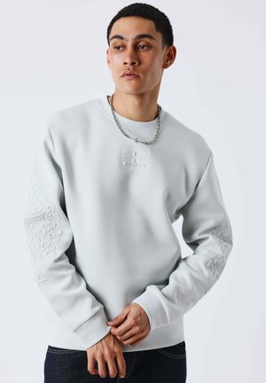 Junger Mann trägt ein hellgraues Sweatshirt mit erhabenen Designs an Ärmeln und Brust sowie eine silberne Kettenhalskette vor einfachem Hintergrund.