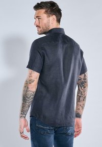 Kurz�ärmeliges, dunkelgraues Leinenhemd mit Kragen, mit abgerundetem Saum und lockerem Schnitt, getragen mit blauen Jeans und sichtbaren Tattoos.